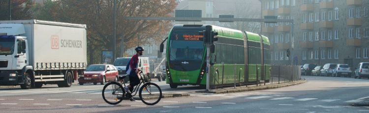 Cyklist som cyklar över större väg, grön buss och lastbil har stannat för att släppa förbi cyklisten.