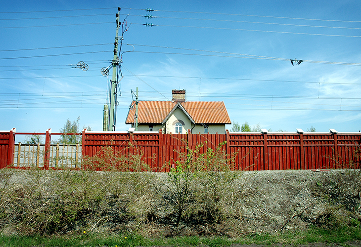 Ett rött bullerplank av trä. Bakom planket syns kontaktledningar till järnväg och ett tegeltak till ett cremevitt hus.