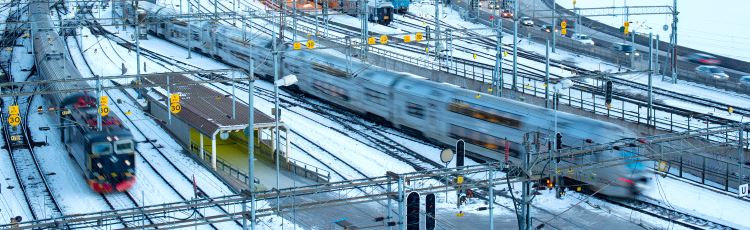 Sex parallella järnvägsspår med kontaktledningsbryggor över spåren, vid infart till större station. Två persontåg kör på olika spår, snö ligger mellan spåren.