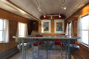 En vagn med träfärgade väggar, två tavlor och ett högt, avlångt bord med grå pallar runtomkring.