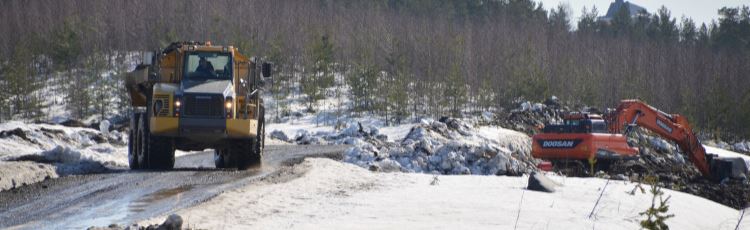 Maskiner som gräver och kör grus på skogsväg, smältande snö på sidorna av vägen. 