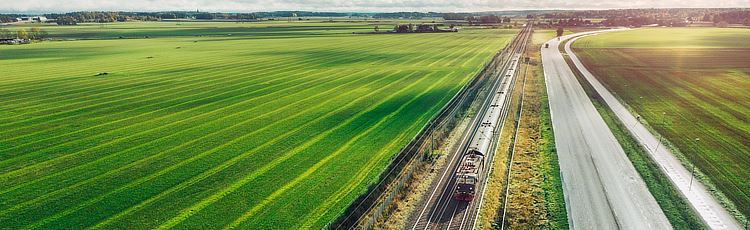 Grönt landskap med väg och järnväg bredvid varandra. Ett tåg åker på spåret.